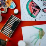 Creative art setup featuring watercolors and pastels on a red table, showcasing artistic expression.
